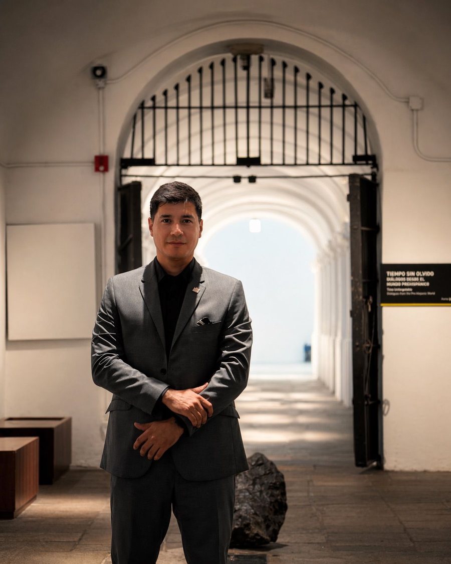 Professional portrait of Andrés Arcila Carmona in a tailored grey suit, standing in an arched museum corridor
